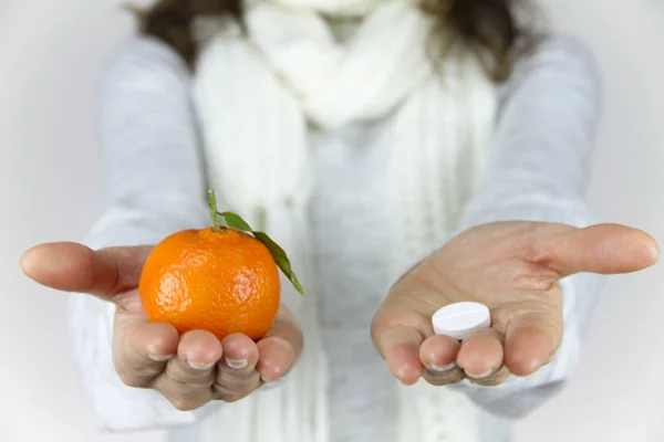 Vitamins from fruit or drugs? A sick young woman with a scarf on her neck shows a mandarin in her right hand and an aspirin on her left