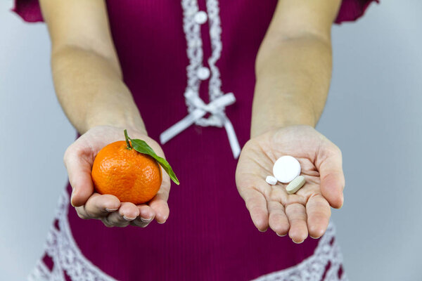 Vitamins from fruits or medicines? A young woman in burgundy pajamas shows a mandarin in her right hand and an aspirin in her left