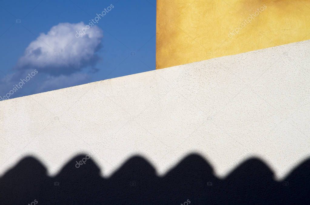 Trapped cloud. Two exterior walls of the house, white and yellow, bound a small piece of blue sky where a small cloud flies. Below the menacing and black shadow of the cornice weighs the composition balancing it