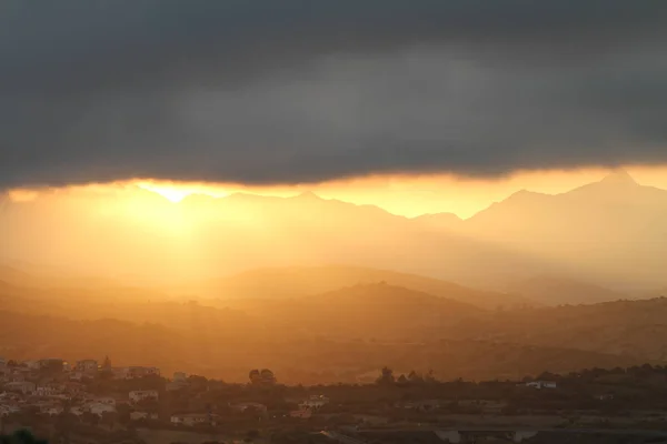 Breathtaking sunset after the storm. The orange sun's rays slip under a big black cloud illuminating the mountains and the hills with the houses of a small village