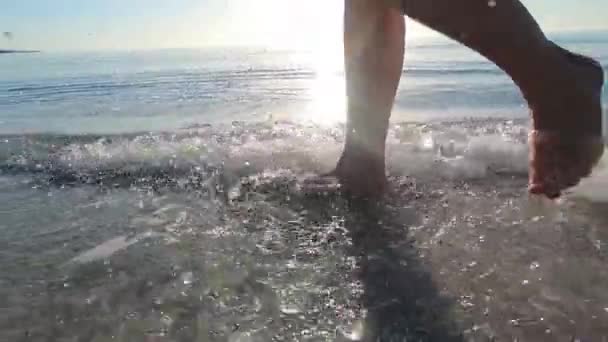一位年轻女子的腿慢慢地沿着海岸走着 海浪冲刷着她的脚 在黎明时分 沙滩上的水花在阳光的映衬下 前景一片光明 — 图库视频影像