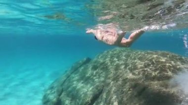 Kayalar ve renkli deniz bitki örtüsü ve beyaz kum deniz dibi üzerinde maske ve şnorkelli yüzme ile mavi suda sualtı yüzmeye alınan siyah bir bikini güzel bir genç kadının yavaş hareket