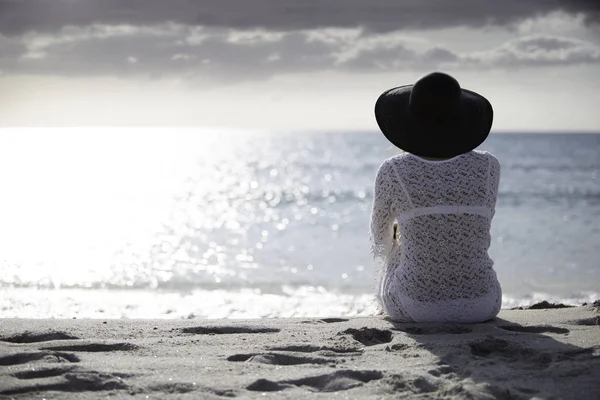 Young woman with long hair from behind sitting by the sea looks at the horizon at dawn in the wind, dressed in a white lace dress, white underwear and large black hat — Stock Photo, Image