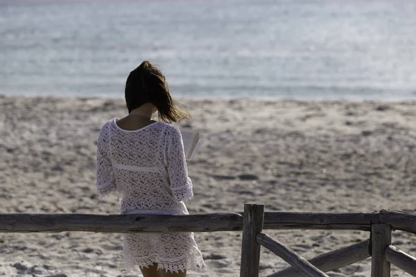 Young woman from behind reads a book on the beach looking at the horizon at dawn in the wind, dressed in a white lace dress, white underwear and long hair — Stock Photo, Image