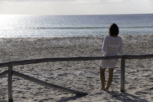 Young woman from behind reads a book on the beach looking at the horizon at dawn in the wind, dressed in a white lace dress, white underwear and long hair — Stock Photo, Image