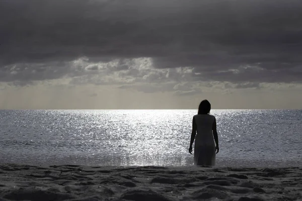 Woman silhouette from behind dressed in a light white dress walks sadly and thoughtfully towards the dark sea at dawn with dramatic sky — Stock Photo, Image