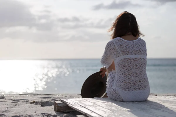 Young woman from behind sitting by the sea looks at the horizon at dawn in the wind, dressed in a white lace dress and white underwear and long hair — Stock Photo, Image