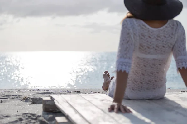 Young woman with long hair from behind sitting by the sea looks at the horizon at dawn in the wind, dressed in a white lace dress, white underwear and large black hat — Stock Photo, Image