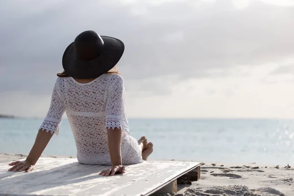 Young woman with long hair from behind sitting by the sea looks at the horizon at dawn in the wind, dressed in a white lace dress, white underwear and large black hat