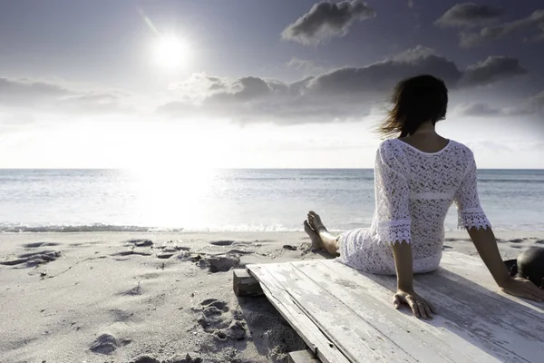 Young woman from behind sitting by the sea looks at the horizon at dawn in the wind, dressed in a white lace dress and white underwear and long hair — Stock Photo, Image