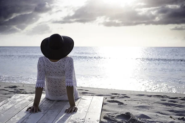 Young woman with long hair from behind sitting by the sea looks at the horizon at dawn in the wind, dressed in a white lace dress, white underwear and large black hat — Stock Photo, Image