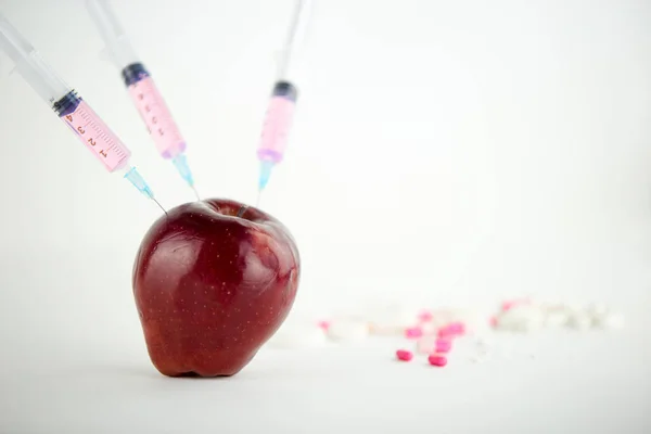 Concept: human GMO manipulation of nature and relative poisoned fruits. Close-up of an apple contaminated with three syringe threaded in and medicines on a white background