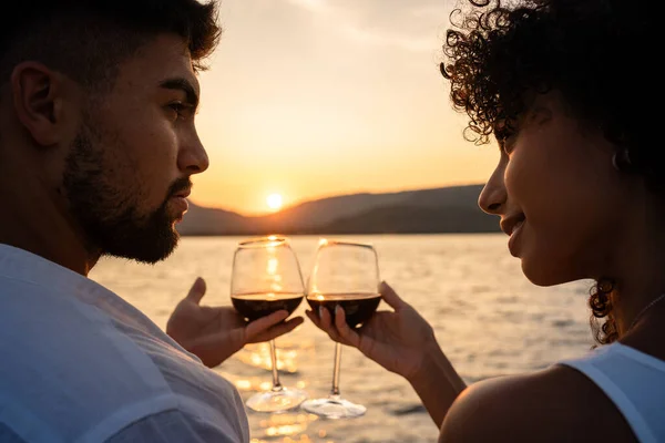 Melez bir çiftin romantik yakın çekimi gölde batan güneşin ışığıyla kesişen kırmızı şarap kadehleriyle.