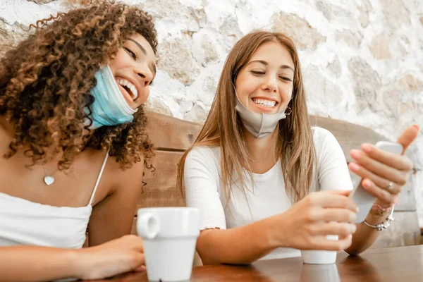 Coronavirus koruma maskeli çok ırklı bir kadın, elinde kahve fincanı olan akıllı telefona bakıp gülüyor. Sarışın en iyi arkadaşıyla gülümseyen İspanyol, tatlı, genç bir kadın.