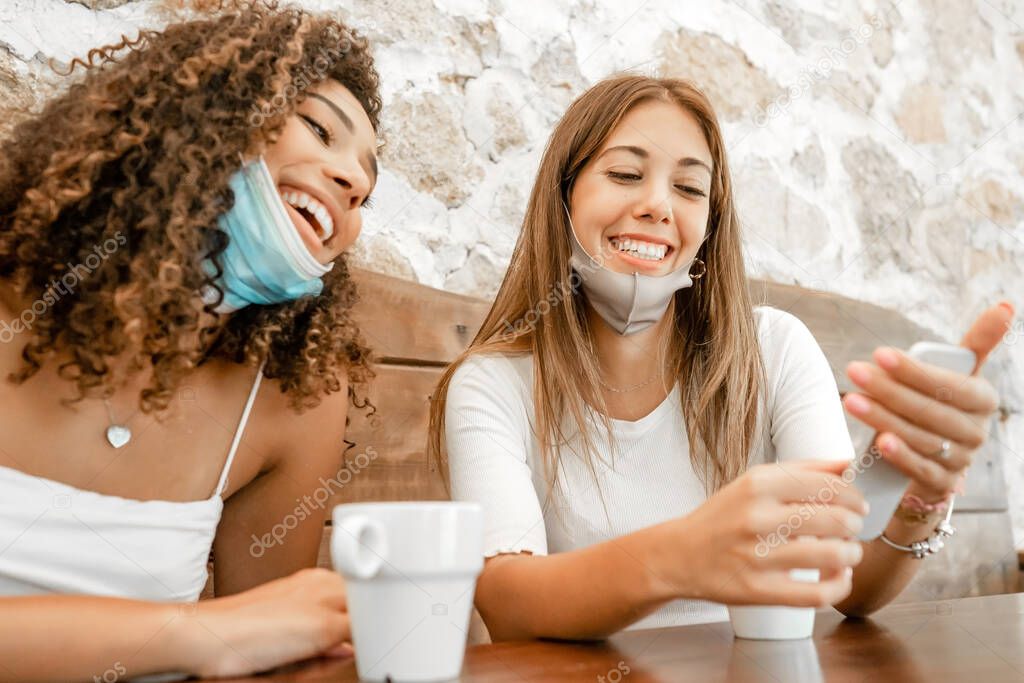 Multi-ethnic female couple with Coronavirus protection mask laughing looking at the smartphone sitting at a table with a coffee cup - Hispanic cute young woman smiling with her blonde best friend