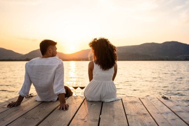 İki kırmızı şarap kadehiyle bir iskelede oturan çok ırklı genç bir çift ve İspanyol. Ve aralarına güneş ışığı koyuyorlar. Romantik günbatımı sahnesi.