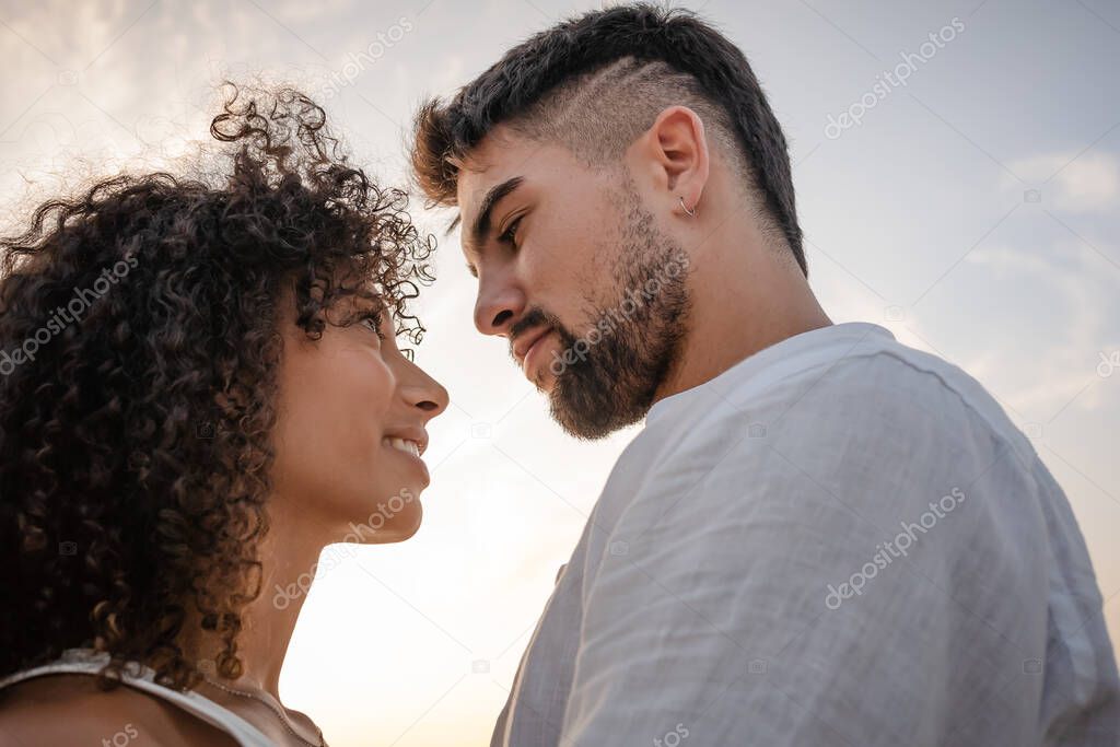 Side view of two young multiethnic passionate lovers looking in the eyes each other - Macho bearded sexy man looks at his Latin woman with ardor in a romance sunset scene ideal for a book cover