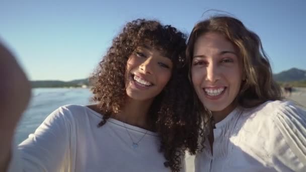 两名年轻貌美的女子一边在海滨度假一边拥抱 一边在海滩上进行视频拜访 日落或黎明时分 在海滩上举行多种族女性情侣视频会议 — 图库视频影像