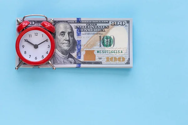 Budzik stoi na stos pieniędzy na niebieskim tle — Zdjęcie stockowe