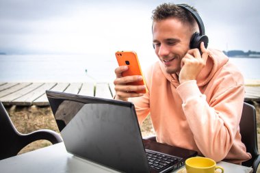 Kafe terasında oturan genç bir adam, kulaklıklı bir dizüstü bilgisayar kullanıyor..