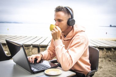 Kafe terasında oturan genç bir adam, kulaklıklı bir dizüstü bilgisayar kullanıyor..