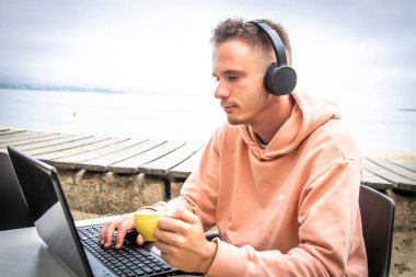 Kafe terasında oturan genç bir adam, kulaklıklı bir dizüstü bilgisayar kullanıyor..