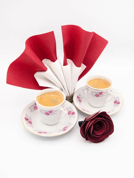 Two floral ornamented porcelain cups of espresso coffee and a paper napkin in a round silver napkin holder decorated with a red paper rose, studio isolated on a white background