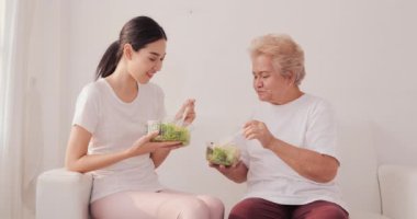 Genç bir kadın ve annesi beyaz koltukta otururken salata yiyorlar.