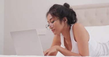 Gözlüklü güzel Asyalı kadın yatakta yatarken dizüstü bilgisayar kullanıyor.