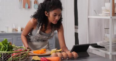 Güzel bir kadın şık bir mutfakta yemek pişiriyor ve tablet bilgisayar kullanıyor.
