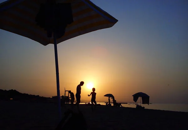 Siluetas de hombre y niño con sombrilla en la playa al atardecer . — Foto de Stock
