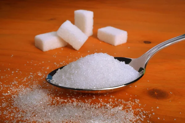 Une cuillère pleine de sucre blanc raffiné sur une table en bois et des cubes de sucre sur fond .. — Photo