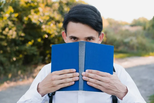 The schoolboy holds a book and looks out from behind it.