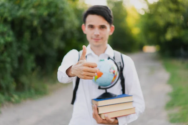 The guy holds a book with a globe in his hand and shows a thumb up on the background of nature.