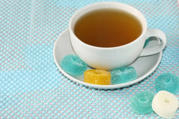 White cup with green tea on a saucer with multi-colored marmalade sweets.Sugar sweet Christmas sweets. Different gelatinous sweet fruit candies for children. Mint, white, yellow, blue background color — Stock Photo, Image