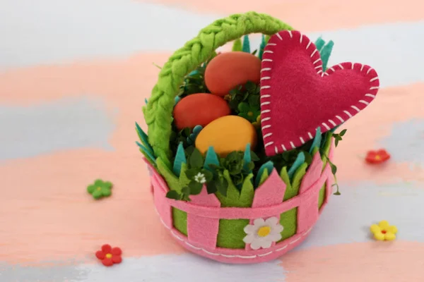 Una cesta de fieltro de colores brillantes con huevos pintados y un corazón rosa sobre un fondo rosa y azul claro. Hecho a mano de fieltro. Feliz Pascua tarjeta de felicitación patrón de fondo. Delicados tonos rosa y azul . — Foto de Stock