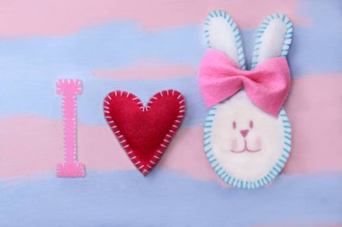 Sevimli beyaz Paskalya tavşanı keçe çok renkli çiçekli pembe-mavi arka plan üzerinde pembe bir yay ile yapılmış. El yapımı keçe üzerinden. Kartpostal mutlu Paskalya için tebrik arka plan. Paskalya süsleri.