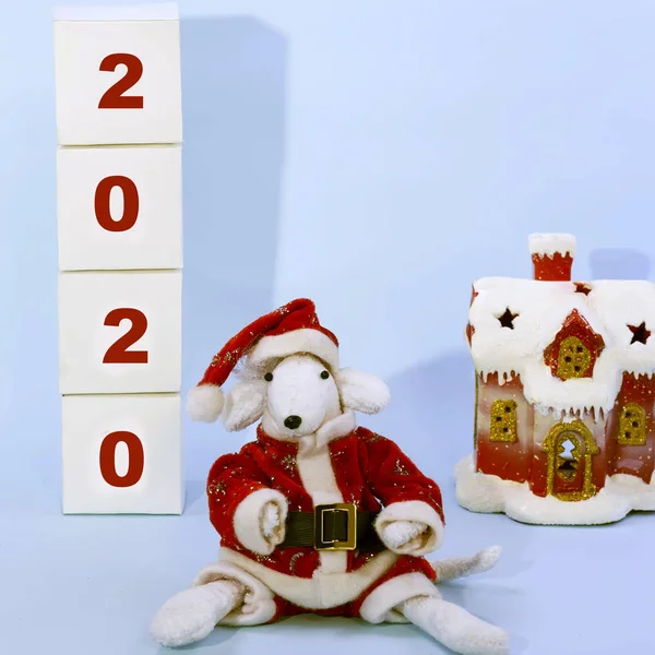 Cute white rat in a red Santa Claus suit in the center of the frame near a snow-covered house and cubes with numbers 2020 on a blue background. Symbol of 2020 on the eastern calendar. — Stock Photo, Image