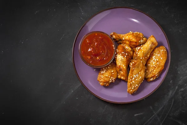 Fried juicy chicken wings marinated with honey, soy sauce, spices, sprinkled with sesame seeds on a plate on a black background with chili sauce. Asian recipe, top view — Stock Photo, Image