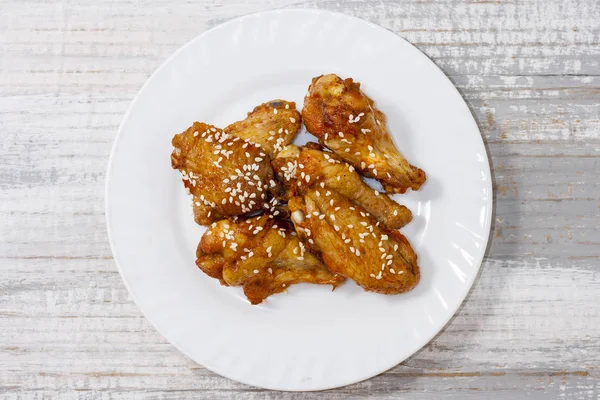 Τηγανητά ζουμερά φτερά κοτόπουλου μαριναρισμένα με μέλι, σάλτσα σόγιας, μπαχαρικά, ψεκάζονται με σουσάμι σε λευκό πιάτο σε ανοιχτόχρωμο φόντο. Ασιατική συνταγή, κορυφή θέα, κοντινό — Φωτογραφία Αρχείου