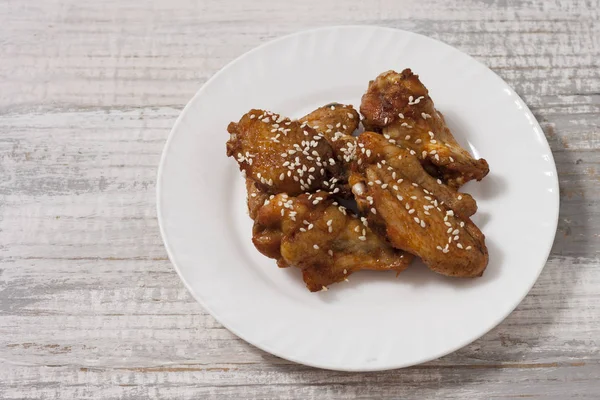 Ali di pollo fritte e succose marinate con miele, salsa di soia, spezie, cosparse di semi di sesamo su un piatto bianco su uno sfondo leggero. Ricetta asiatica, vista dall'alto, primo piano — Foto Stock