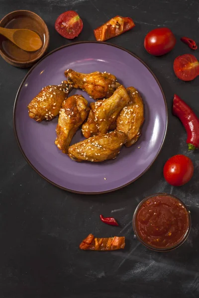 Gebakken sappige kippenvleugels gemarineerd met honing, sojasaus, specerijen, bestrookt met sesamzaden op een bord op een zwarte achtergrond met tomaten, rode peper, honing en Chili saus. Aziatisch recept, bovenaanzicht — Stockfoto