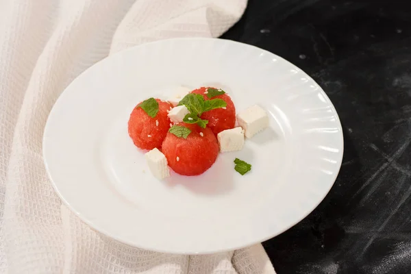 Salat aus Wassermelonenbällchen, Feta-Käse und Minze, bestreut mit Sesam in einem weißen Teller auf schwarzem Hintergrund. — Stockfoto