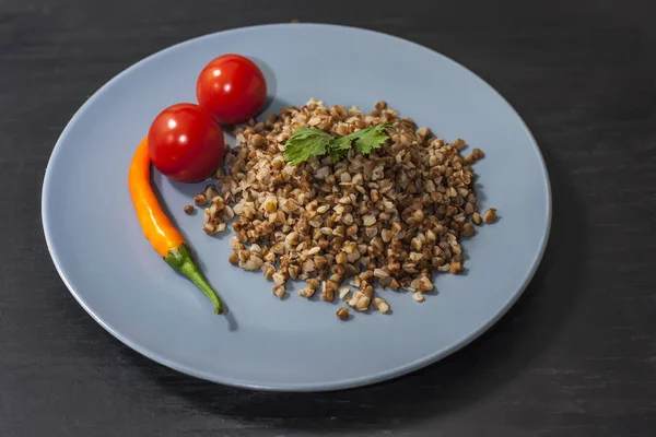 Mingau de trigo mourisco cozido em uma placa cinza com legumes em um fundo de madeira preta  . — Fotografia de Stock