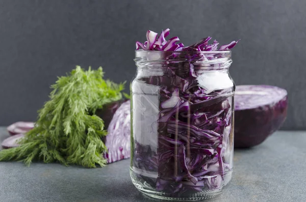 Glass jar with chopped fresh red cabbage,dill and rings of onion- preparation for fermentation on the grey surface in the kitchen.