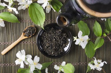 Yeşil hasır mat üzerinde kuru yeşil çay, ahşap kaşık ve aromatik taze yasemin çiçekleri dolu siyah Çaydanlık ve daire üst görünümü