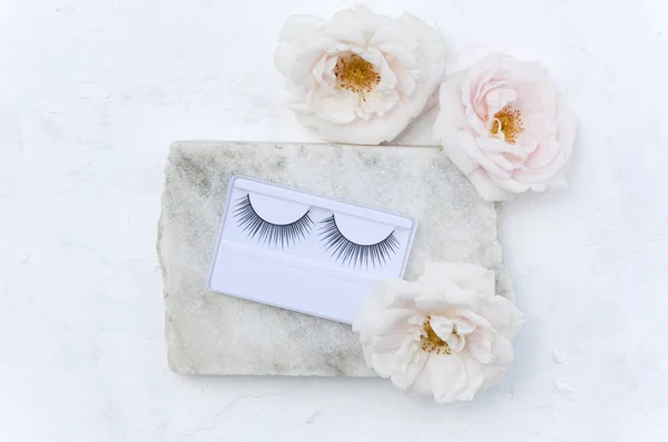 Closeup of false eyelashes and beautiful flowers on the piece of marble stone on the white surface.Beauty concept