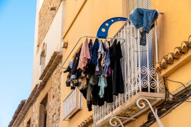 Denia, Alicante - İspanya - 12-22-2023: Temiz bir gökyüzü altında eski bir binanın ön cephesine karşı balkonda kuruyan elbiseler