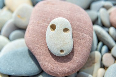 A stone with three natural perforations lies on a reddish rock, resembling a face, set against a pebble backdrop clipart