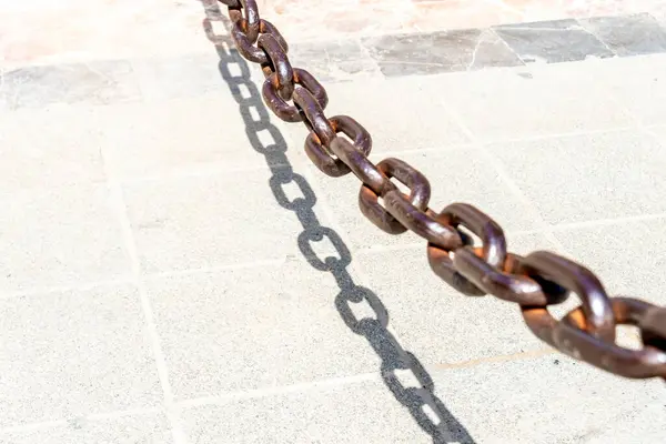 Rusty brown chain casting a sharp shadow on sunlit paving stones